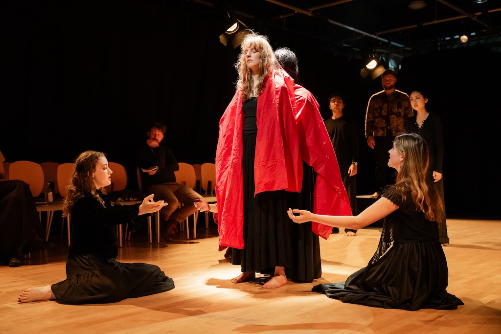 Two actors wearing bright red capes stand with their backs to one another while other actors kneel before them with their arms outstretched.